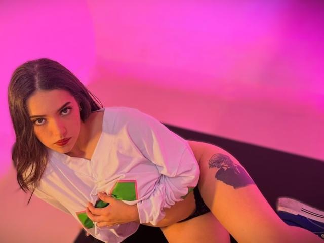 KatarinaNovak with wet brown hair, white shirt, black panties, holding green card, leaning on black surface, visible tattoo on right thigh, pink and purple lighting, looking at camera.