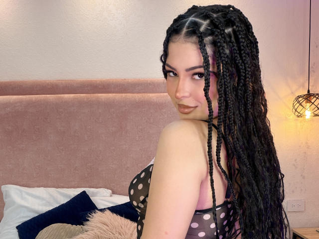 RheaVolk with long black braids, wearing a sheer black polka dot top, showing her tits, looking over her shoulder, in a bedroom with pink headboard and warm light.