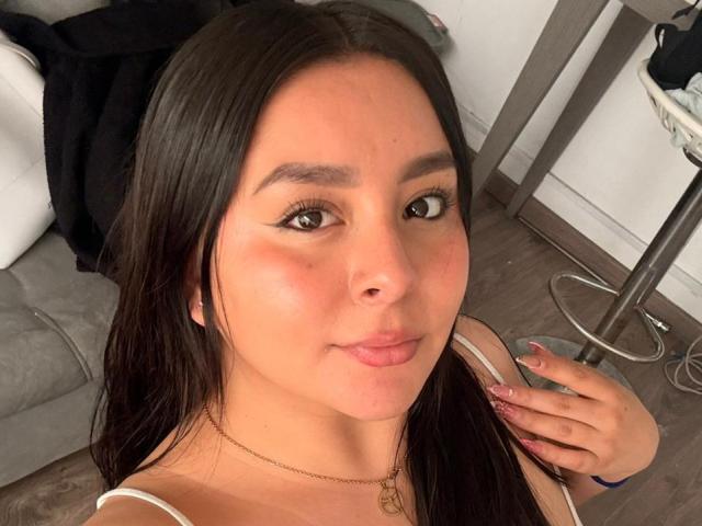EmaJames has brown eyes, long dark hair, and is wearing a white top with a gold necklace. She's pouting slightly with a hand near her chin.