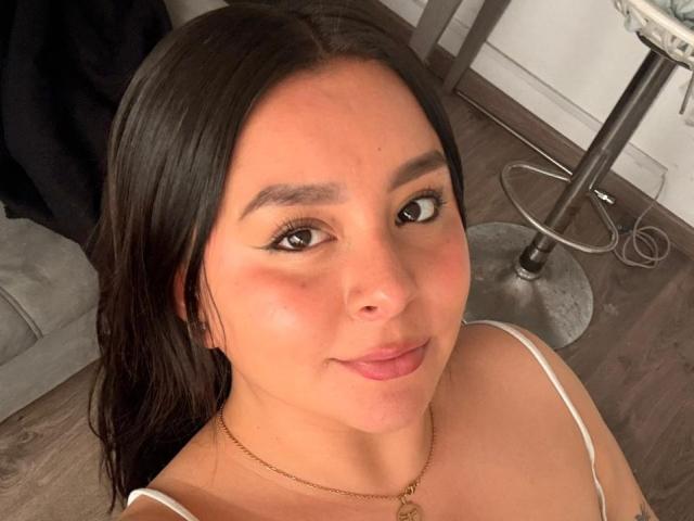 EmaJames with dark, straight hair, brown eyes, subtle makeup, pink lips, wearing a white top, gold necklace, and slight blush, looking up at the camera with a slight smile.