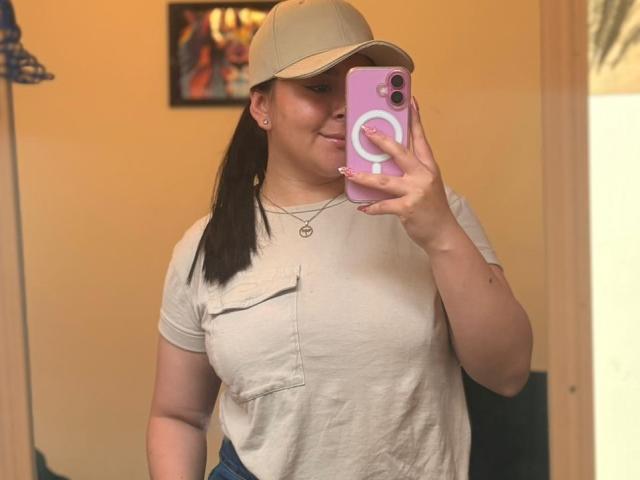 EmaJames with dark ponytail, beige cap, white tee showing her tits, blue jeans, holding pink phone, smiling, wearing necklace, standing in front of mirror, taking selfie.