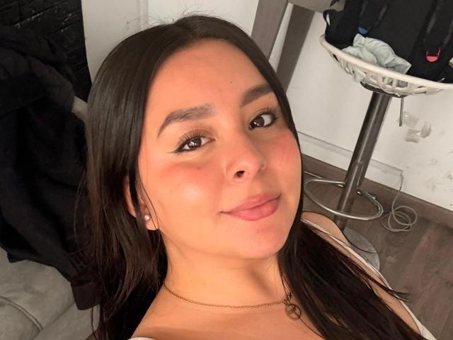 EmaJames with long brown hair, light brown skin, subtle makeup, and a small smile, showing her bare shoulders and a thin gold necklace, in a selfie.