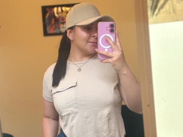 EmaJames with brown ponytail, beige cap, beige shirt showing small tits, holding pink phone, taking mirror selfie, smiling, small earrings, yellow wall, colorful painting in background.