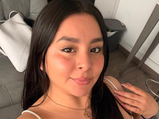 EmaJames has tan skin, dark brown hair, and a subtle smile. She's wearing a white top with a gold necklace, and has a hand with pink nails near her chin.