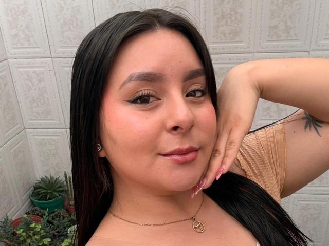 EmaJames with long black hair, wearing a tan top, has a cute smile, raised hand, and a peace symbol necklace, showing a small tattoo on her arm.