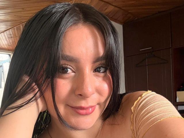 EmaJames with dark straight hair, brown eyes, and a slight smile, wearing a yellow off-shoulder top, showing her cute face and small, round boobs. Wooden ceiling background.