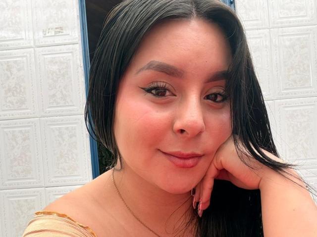EmaJames has dark, wet hair, brown eyes with black eyeliner, a slight smile, and a pinkish blush. She rests her chin on her hand, showing a cute face.