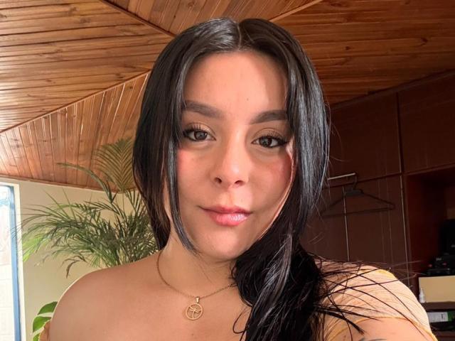 EmaJames has long black hair, brown eyes, and a slight smile. She's wearing a gold necklace with a small pendant. Her bare shoulders are visible. Wood ceiling.