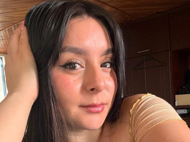 EmaJames with brown eyes, black hair, and pinkish skin, holding her head with her hand, wearing a yellow top, showing her slightly shiny pink lips.