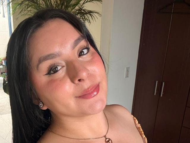 EmaJames has shiny brown eyes, black hair, and a subtle smile. She's wearing a necklace with a small heart, and has cute earrings. Her tits are slightly visible.