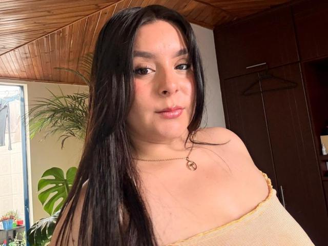 EmaJames has long black hair, a gold necklace, and a yellow off-shoulder top showing her tits. She's looking at the camera with a slight smile.
