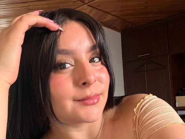 EmaJames has dark hair, brown eyes, and pinkish lips, wearing a yellow top with ruffled shoulders, her hand in her hair, smiling slightly, in a wooden ceiling room.