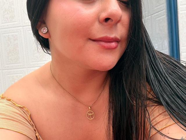 EmaJames with long black hair, light skin, and pink lips, wearing a gold necklace with a circle pendant, off-shoulder top, and pink earrings, smiling slightly.