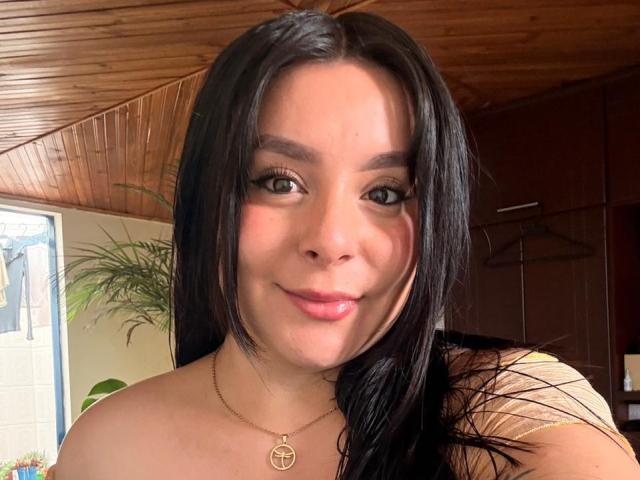 EmaJames has long black hair, brown eyes, and a slight smile with pink lips. She's wearing a gold necklace with a pendant and a yellow top.