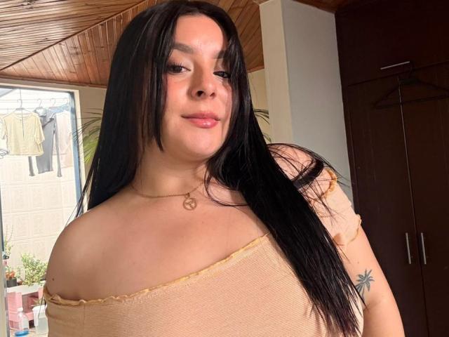 EmaJames with long black hair, light skin, and small tits in a beige off-shoulder top, smiling slightly, wearing a heart necklace, standing near dark wood wardrobe.