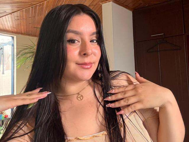 EmaJames with long black hair, pink lipstick, and a subtle smile, wearing a yellow top, hands near face, pink nails, peace necklace, wooden ceiling, and a dark brown door.