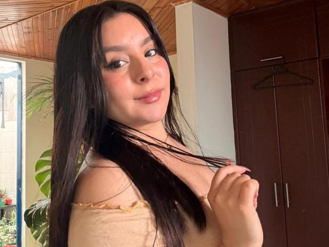 EmaJames with long black hair, slightly smiling, holding her hair, wearing a beige off-shoulder top, brown wooden wardrobe in background.