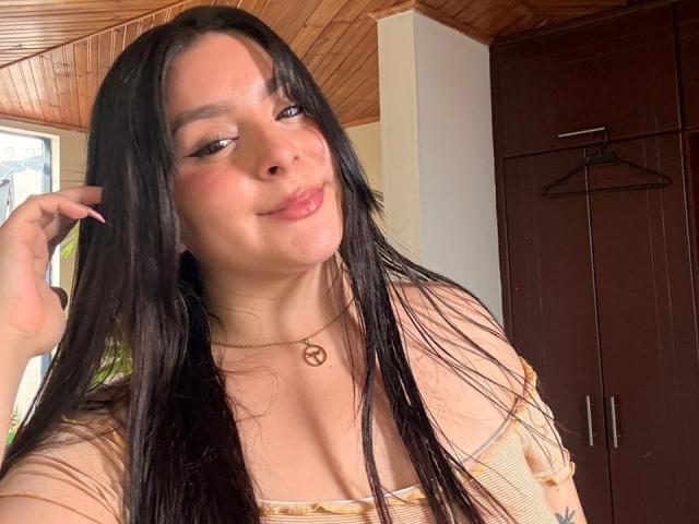 EmaJames with long black hair, wearing a gold necklace with a peace sign, slightly smiling, showing her tits in a sheer top, holding her hair.