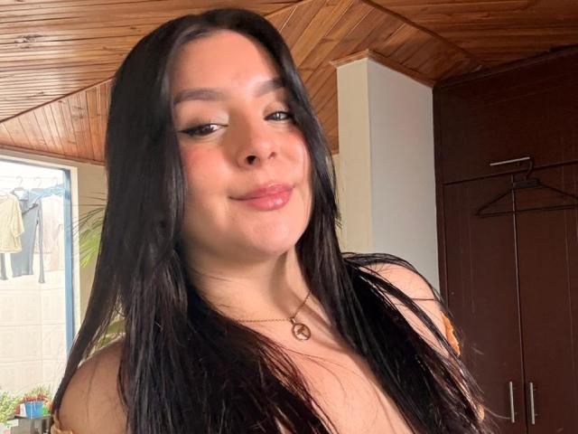 EmaJames with long black hair, light brown skin, and subtle makeup, wearing a necklace with a small circular pendant, has her tits slightly visible, smiling at the camera.
