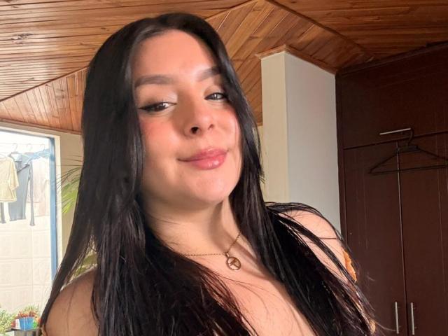EmaJames with long black hair, brown eyes, and a small smile, wearing a necklace, has shiny pink lips and is showing off her tits in a wooden ceiling room.