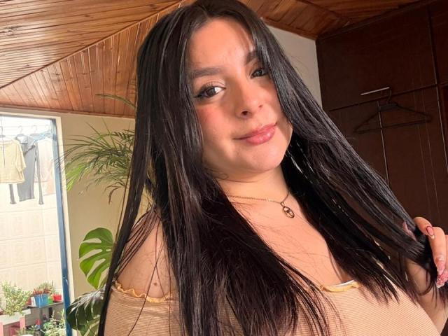 EmaJames has long black hair, light brown skin, and is smiling with a cute nose. She's wearing a beige top, showing her small tits, and holding her hair.
