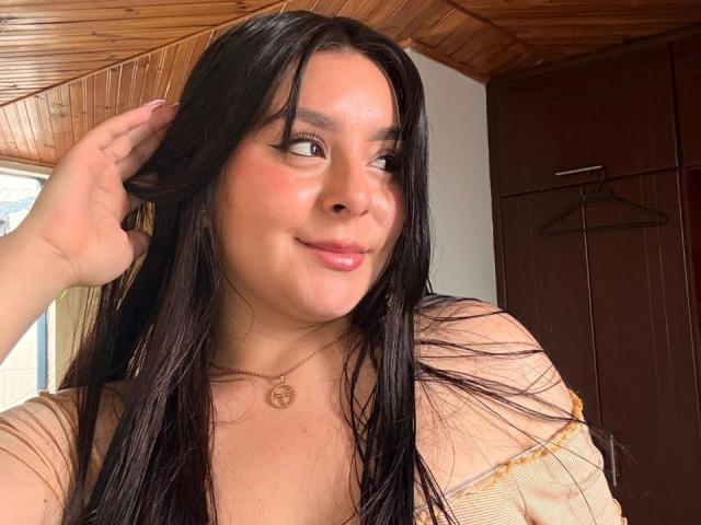 EmaJames has long black hair, a cute smile, and is touching her head with one hand. She's wearing a sheer top, showing her tits, and a gold necklace.