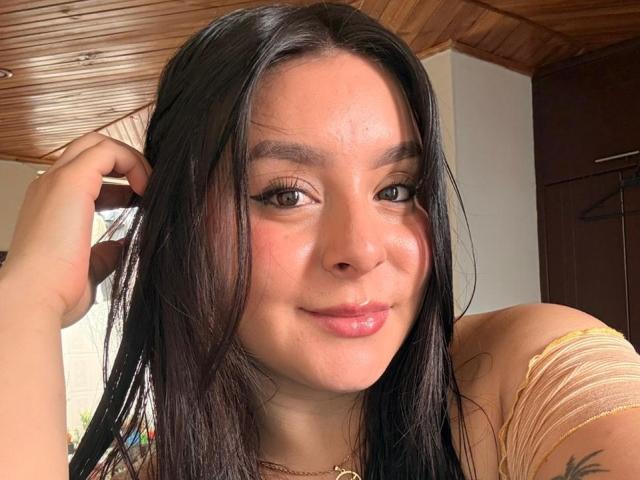 EmaJames with long black hair, light skin, and brown eyes, slightly glossy pink lips, wearing a yellow top, touching her hair, wooden ceiling, and brown door in background.