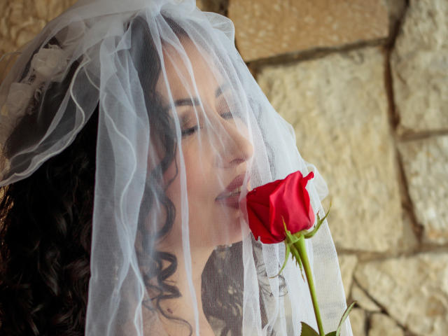 SamanthaAntolin with curly black hair, wearing a white veil, gently kissing a red rose, eyes closed, soft smile, stone wall background, holding rose with green stem.