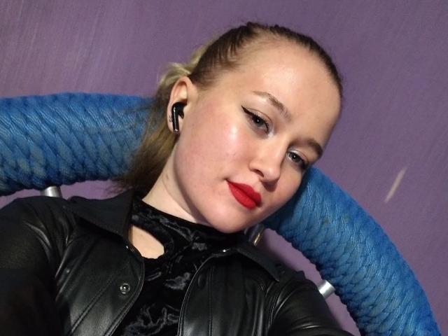 Claire Meyerman, met rode lippenstift, een zwarte leren jas en blauwe oordopjes, draagt een zwarte kanten top, heeft blond haar in een paardenstaart, en er staat een blauwe krullenstoel achter haar. Op de achtergrond is een paarse muur te zien..