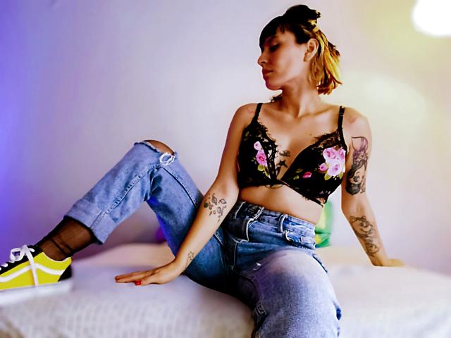 SaraleePoison with tattoos, wearing a black floral bra, ripped jeans, and neon yellow sneakers, sitting on a bed, looking to the side, with her ass up.