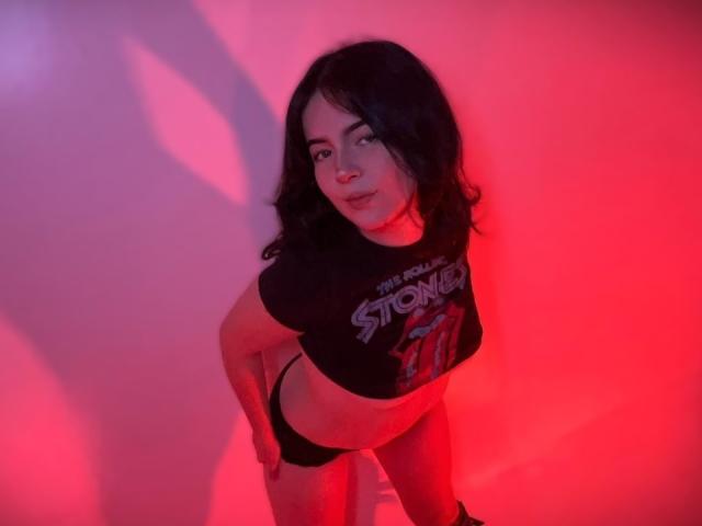 AliinaFontaine mit schwarzen Haaren, die ein schwarzes Crop-Top und schwarze Unterwäsche trägt, lehnt sich nach vorne und hat die Hände auf den Hüften, unter rotem und rosa Licht, wobei ihr Hintern zu sehen ist..