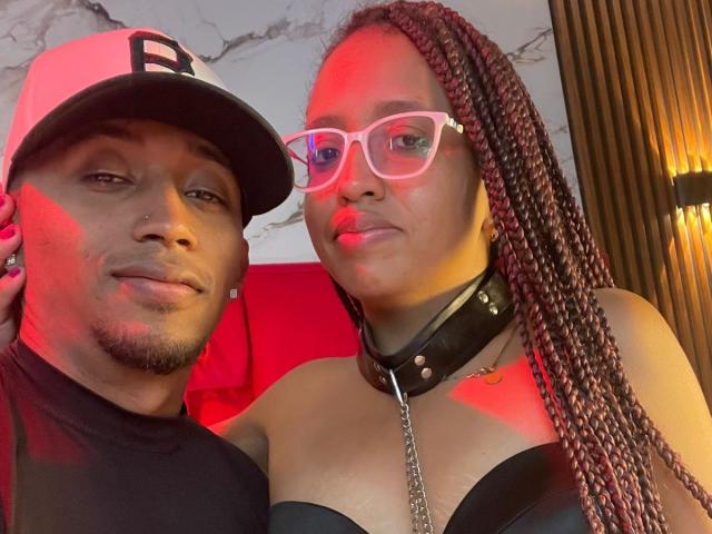 LilithAndAsmodeo with braided red hair, pink glasses, black leather strap top, collar with chain, and LilithAndAsmodeo in black shirt, cap, and nose ring, both with red lighting.