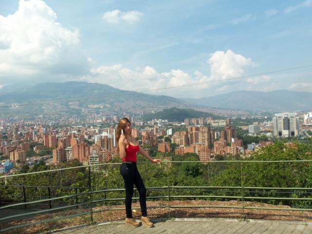 HotPamelaSex stands on a concrete ledge, wearing a red top, black pants, and tan boots, with her brown hair in a ponytail, looking forward, in a cityscape view.