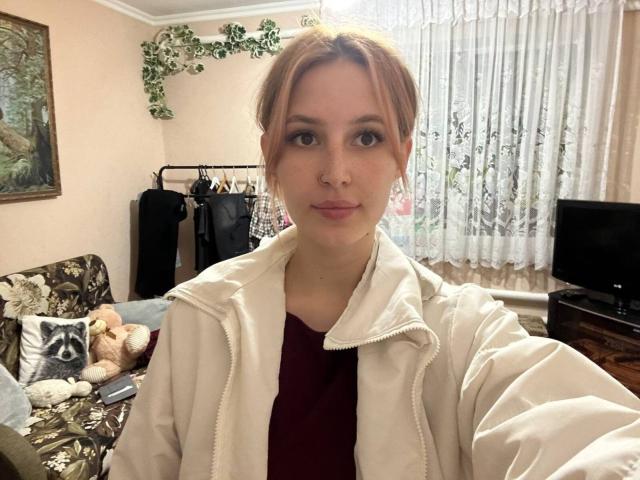 Brynn Haynesworth, de piel pálida, con cabello castaño claro recogido en un moño, vestida con una chaqueta blanca sobre una blusa color burdeos, mirando a la cámara con una ligera sonrisa, sosteniendo un palo para selfies..