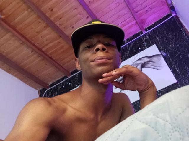 MasterXx, shirtless, dark-skinned, wearing black cap with yellow brim, slightly smirking, hand resting on chin, wooden ceiling, purple and white lighting, black marble wall, gray quilt.