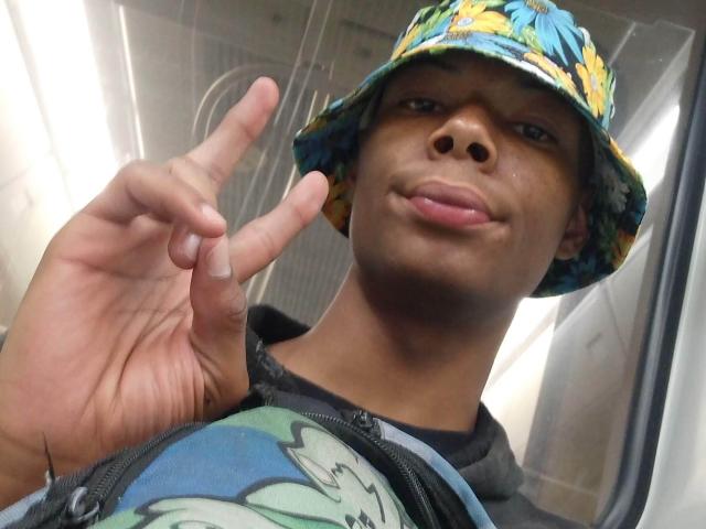 MasterXx with dark skin, wearing a colorful floral bucket hat, black hoodie, and a graphic tee, making a peace sign, with a slight smirk, and glossy lips.