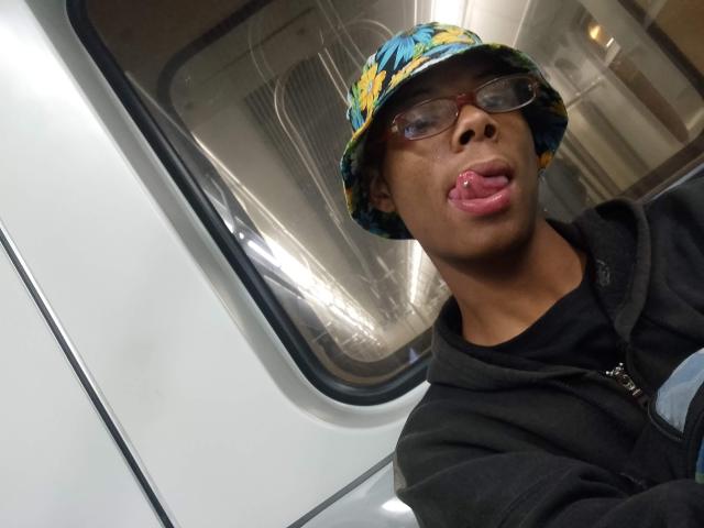 MasterXx, with dark skin, wearing a floral bucket hat and glasses, sticking out their pink tongue, in a black hoodie, standing by a white train window.