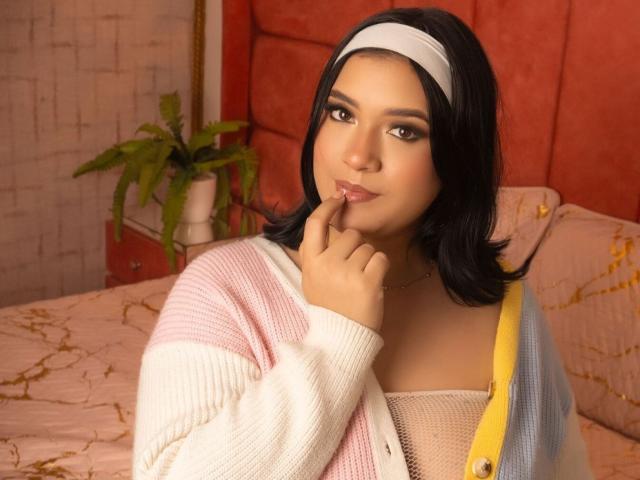 NathalyParker has black hair, a white headband, and a pink-and-white cardigan, with a finger on her chin, looking cute and confident, showing her soft, rounded tits.