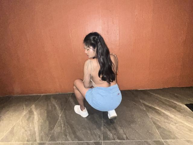 EsaMarie, squatting, topless, showing her ass, wearing a blue skirt, white shoes, long black hair, red textured wall, brown tiled floor, looking over her shoulder.
