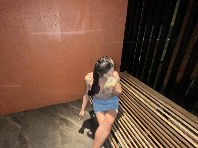 EsaMarie with long brown hair, tan tank top, and blue skirt, sitting on a wooden bench, drinking from a yellow cup, holding her phone, looking down.
