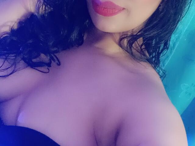 FetishLoverAna's face with red lipstick, curly black hair, and large, bare boobs, slightly covered by a black strap. Soft blue lighting.