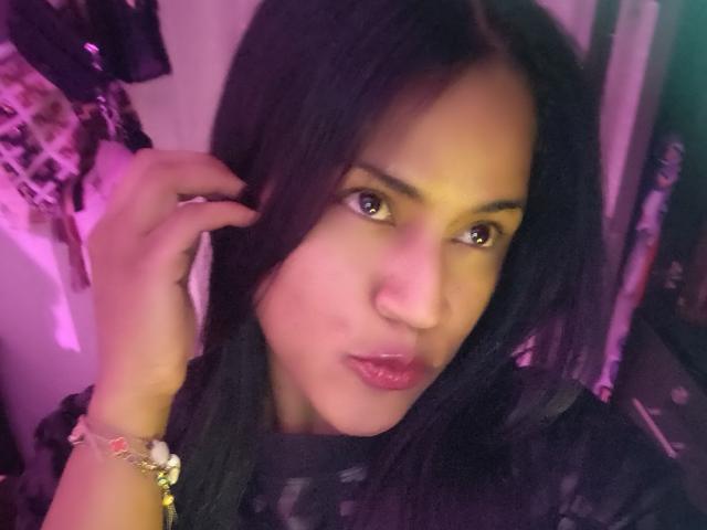 IvannaDuran with dark, straight hair, brown eyes, pink lips, and a bracelet on her left wrist, holding her hair with a slight pout. Purple lighting.