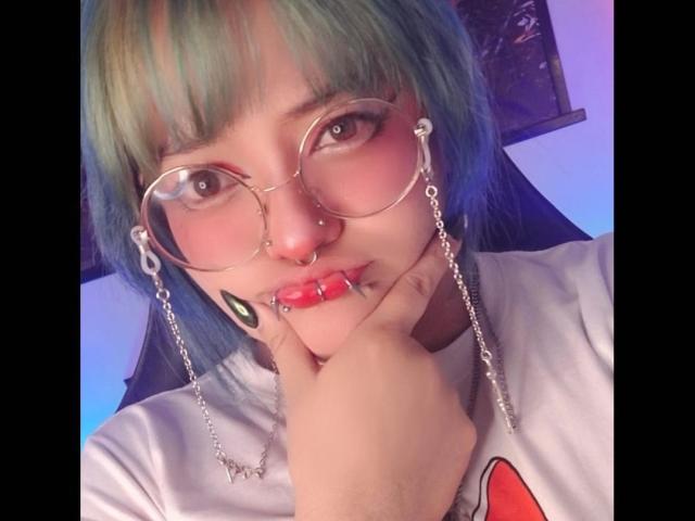 LuckyLeahh, con i capelli blu-verdi, gli occhiali rotondi, il piercing al naso, il piercing al labbro e gli orecchini a catena, che tocca le sue labbra rosa con un dito, indossa una camicia bianca e sembra adorabile..