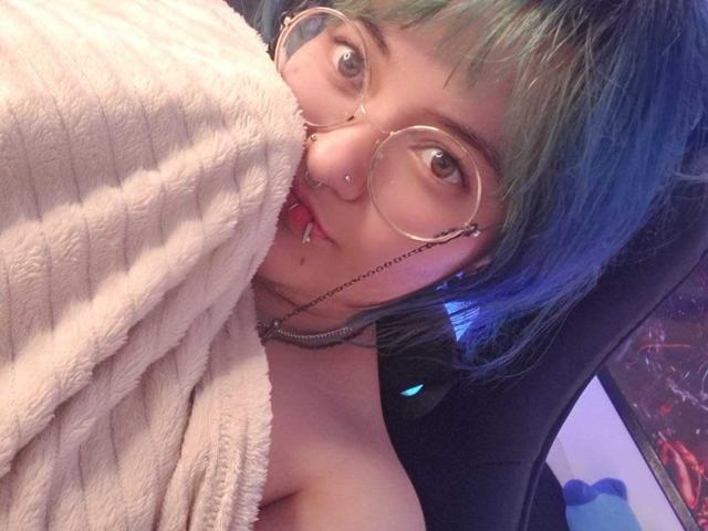 LuckyLeahh with blue-green hair, glasses, nose ring, and lip piercing, wrapped in a fluffy beige blanket, showing bare shoulders, sitting in a chair, looking at camera.