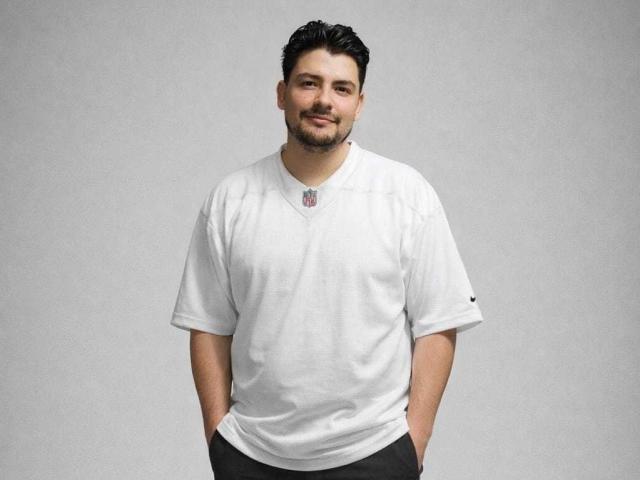 DracoSeoul, ein etwas korpulenter Mann mit kurzen, dunklen Haaren und Bart, trägt ein weißes T-Shirt mit V-Ausschnitt, schwarze Hosen, steht da und lächelt leicht..