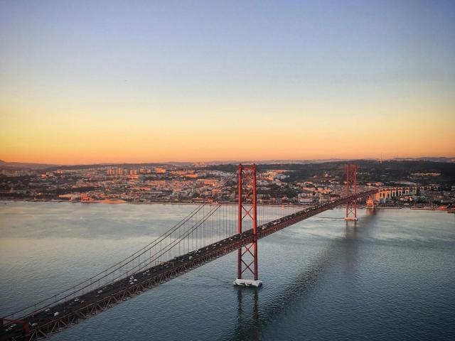 Vista aérea de un puente colgante rojo con coches, que se refleja en agua tranquila, bajo un cielo claro al atardecer. La ciudad y las colinas de Sunny26-hot se ven en el fondo..