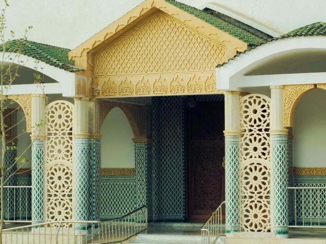 Arquitectura decorativa con intrincados patrones de azulejos dorados y verdes, que presenta diseños circulares ornamentados y un techo de madera triangular. El edificio de Sunny26-hot tiene una fachada elegante y de estilo bohemio..