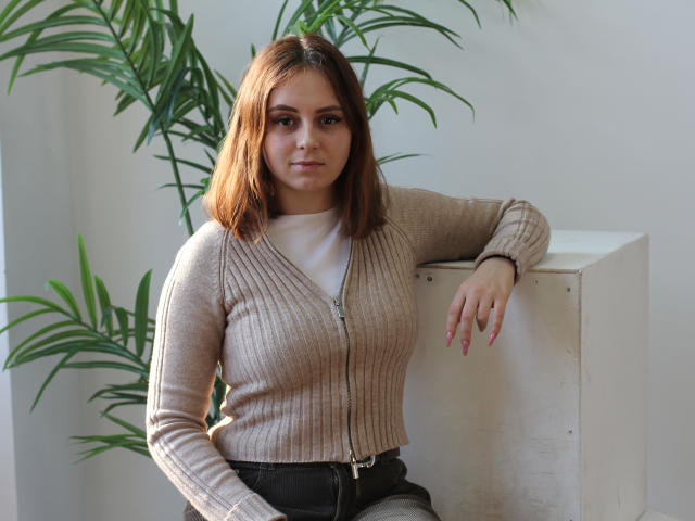 MilanaMiller, brown-haired woman with medium tits, wearing a beige ribbed cardigan and black pants, leans on a white cube, green plants in the background.