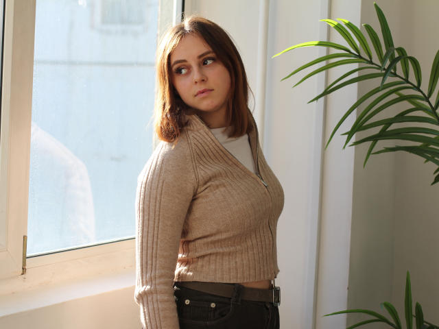 MilanaMiller with medium tits, brown ribbed sweater, and dark jeans, standing by a window, looking to the side with a pensive expression, green plant to the right.