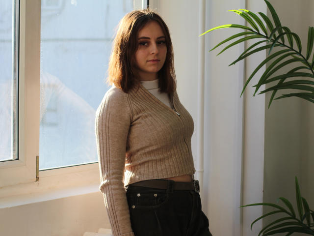 MilanaMiller has brown hair, a serious expression, and wears a beige rib-knit crop top, black high-waisted pants, with a taut belt, standing by a sunlit window.